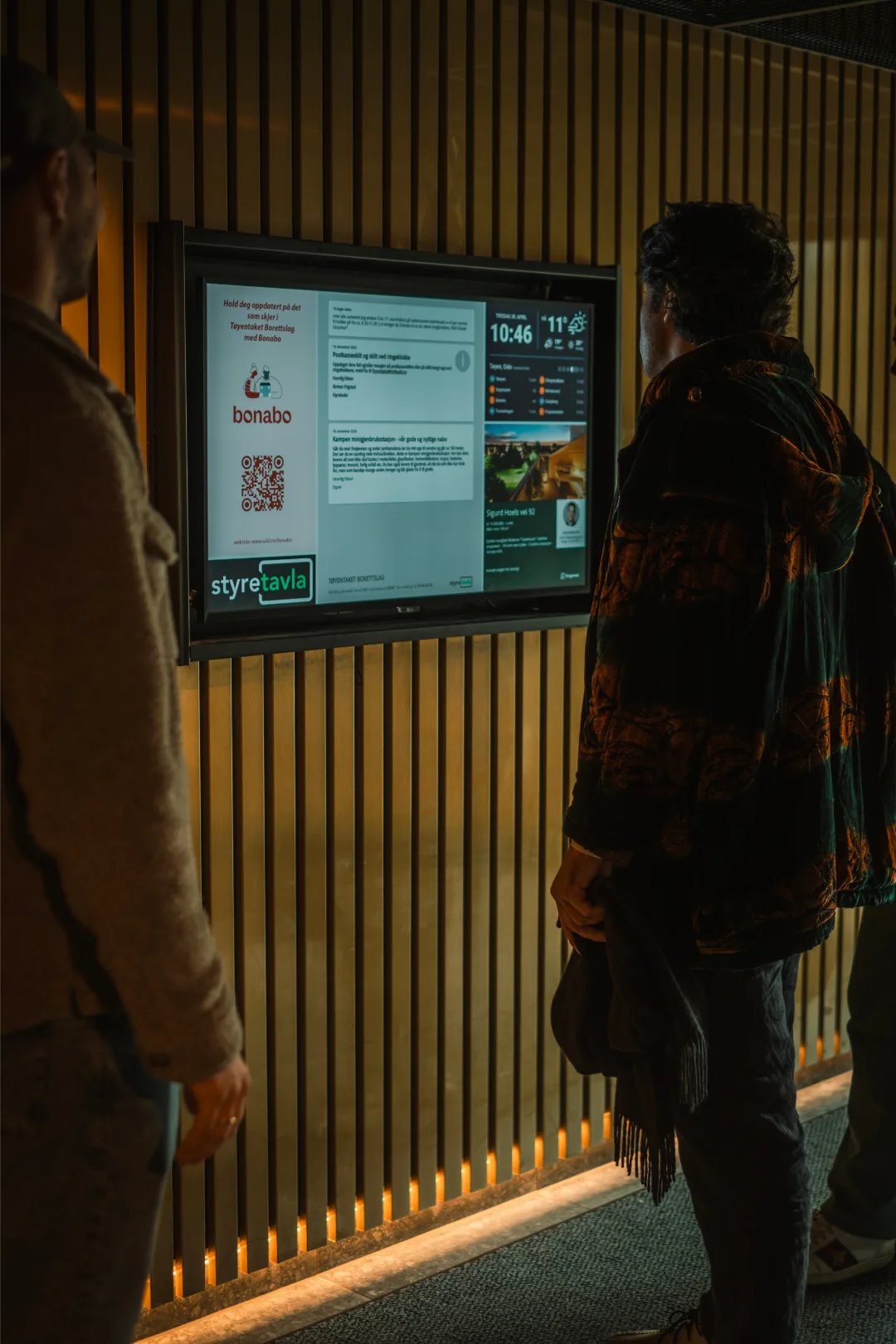 To beboere leser Styretavla-skjermen i en oppgang med trepanel og varmt lys